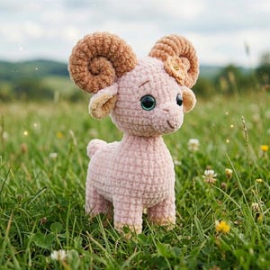 Könnte beinhalten: Handgehäkeltes Widder-Spielzeug aus weichem rosa Garn, mit großen, braunen, lockigen Hörnern und einer kleinen Blume. Der Widder hat große, ausdrucksstarke Augen und steht in einem Feld aus grünem Gras und Wildblumen.