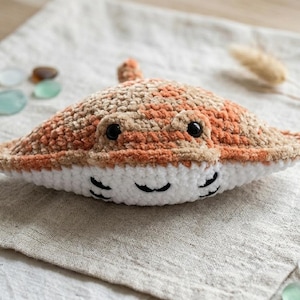 Puede incluir: Un juguete de peluche de manta raya hecho a mano con ganchillo, en tonos naranja, beige y blanco. El peluche tiene ojos negros y una boca sonriente. Descansa sobre una superficie de lino de color neutro, con guijarros de vidrio en el fondo.