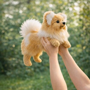 May include: A handmade, plush Pomeranian dog with a fluffy tail and ears. The dog is a mix of tan and white colors, with a detailed crocheted texture. The dog is being held up in the air against a blurred green background.