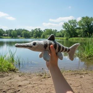 May include: A crocheted ichthyosaur toy in shades of gray and white, featuring a long snout, dorsal fin, and flipper-like appendages. The handmade plush toy is held up against a backdrop of a lake and trees, suggesting an outdoor setting.