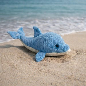 May include: A crocheted blue dolphin toy with a white belly, fins, and tail, resting on a sandy beach. The dolphin has a friendly expression with blue eyes. The background shows the ocean and waves.