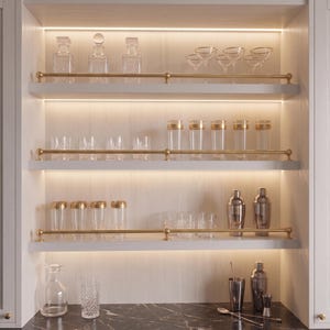 May include: A built-in bar cabinet with three illuminated shelves featuring brass rails. The shelves hold various clear glass decanters, stemmed cocktail glasses, tall tumblers, and metallic cocktail shakers, with additional glassware and bar tools on the counter.