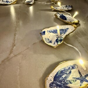 Puede incluir: Una cadena de luces decorativas con conchas de ostra con diseños azules y blancos y bordes dorados. Cada concha contiene una pequeña luz de tono cálido. Las conchas están conectadas por un cable blanco, creando una estética costera única.