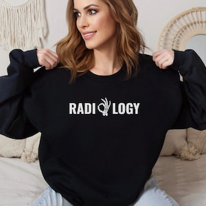 Può includere: Felpa nera con la scritta "RADIOLOGY" in bianco. La "O" nella parola è sostituita da una mano di scheletro bianca che fa il gesto "ok". La felpa è mostrata da una persona.