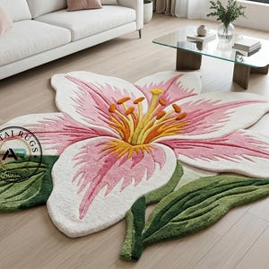 Op de afbeelding: Een groot, bloemvormig tapijt met een lelie-ontwerp. Het tapijt heeft witte, roze, gele en groene kleuren. De lelieblaadjes zijn wit met roze randen en het midden heeft gele en oranje details. Groene bladeren maken het ontwerp compleet.