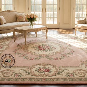 May include: A large, rectangular pink rug with a floral design and a cream-colored border. The rug features a central medallion with roses and leaves, surrounded by a detailed scrollwork pattern. The rug is in a room with antique furniture.