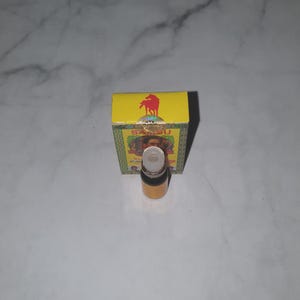 Puede incluir: Una pequeña botella de vidrio transparente con una tapa blanca y un líquido amarillo en su interior. Detrás de la botella hay una caja amarilla con la palabra "Samsu" y un logotipo de caballo rojo. La caja tiene elementos decorativos verdes y dorados.