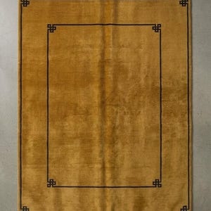 Könnte beinhalten: Ein rechteckiger senfgelber Teppich mit einer dunkelbraunen Umrandung, die an allen vier Ecken mit geometrischen Knotenmustern verziert ist. Der Teppich hat einen kurzflorigen Flor und ist an beiden Enden mit hellen Fransen versehen.