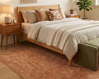 May include: A rectangular area rug in a terracotta orange hue featuring a grid pattern of white stitched crosses. The rug is placed on a light wood floor in a bedroom setting with a wooden bed frame, neutral bedding, and a green bench.