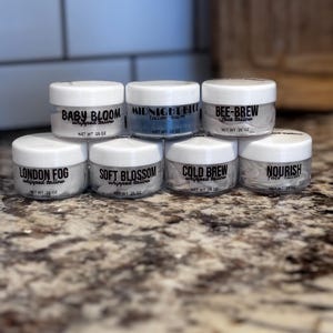 Peut inclure: Plusieurs petits pots à couvercles blancs contenant du baume pour le visage au suif fouetté. Chaque pot porte une étiquette noire avec des noms tels que "Baby Bloom" et "Midnight Blu". Les pots sont disposés sur un comptoir moucheté.