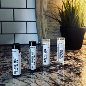 May include: Four lip balm tubes with black and white labels are arranged on a speckled countertop. The flavors are Wedding Mint, Sweet Orange & Mint, Honey-Lemon, and Peppermint. The background includes a cutting board and a potted plant.