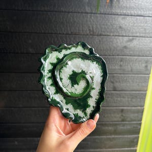 Op de afbeelding: Een keramieken schaal in de vorm van een gegolfd blad met een glanzende afwerking. Het oppervlak heeft een wervelend spiraalpatroon in diep bosgroen en wit, met donkergroene randen en een gestructureerde, organische vorm.