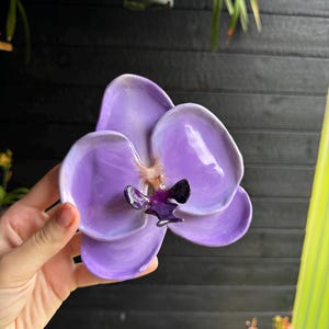 Puede incluir: Una escultura de flor de orquídea de cerámica con pétalos de color lavanda suaves y brillantes, con un centro de color púrpura intenso y marrón oscuro. La flor tiene una forma redondeada y orgánica con un acabado reflectante y color violeta.