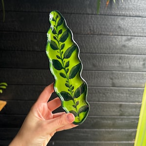 Può includere: Un vassoio in ceramica allungato con bordi ondulati caratterizzato da uno sfondo verde lime vibrante con un motivo botanico a vite verde scuro dipinto a mano al centro, rifinito con uno smalto lucido e bordi dipinti in verde scuro.