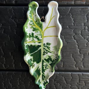 Op de afbeelding: Een keramieken schaal in de vorm van een bontgekleurd blad met een glanzende afwerking. Het oppervlak heeft een geschilderd patroon van witte, lichtgroene en donkergroene vlekken met een opvallende gele hoofdnerf en golvende, geschulpte randen.