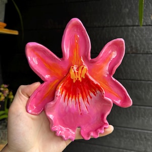 Könnte beinhalten: Eine glänzende Keramikschale in Form einer leuchtend rosa Orchideenblüte. Die Blütenblätter haben eine glatte, glänzende Oberfläche mit bemalten Details in leuchtendem Pink, tiefem Orange und Gelb, die von der Mitte ausstrahlen.