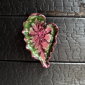Könnte beinhalten: Eine Keramikschale in Form eines herzförmigen Blattes mit glänzender Oberfläche. Die Schale zeigt ein tiefrotes Zentrum mit leuchtend rosa Adern, das in einen dunkelgrünen Rand mit hellrosa und weißen Punkten an den Rändern übergeht.
