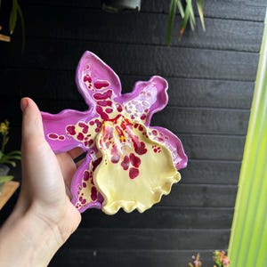 May include: A ceramic dish shaped like an orchid flower with wavy edges. The petals are a vibrant purple with deep red and yellow spotted patterns, while the central labellum is a smooth, pale yellow with dark red speckles and markings.