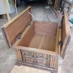 May include: A wooden chest with a hinged lid and three drawers. The chest is made of light brown wood with darker brown accents and metal hardware. The lid is open, revealing the interior of the chest.