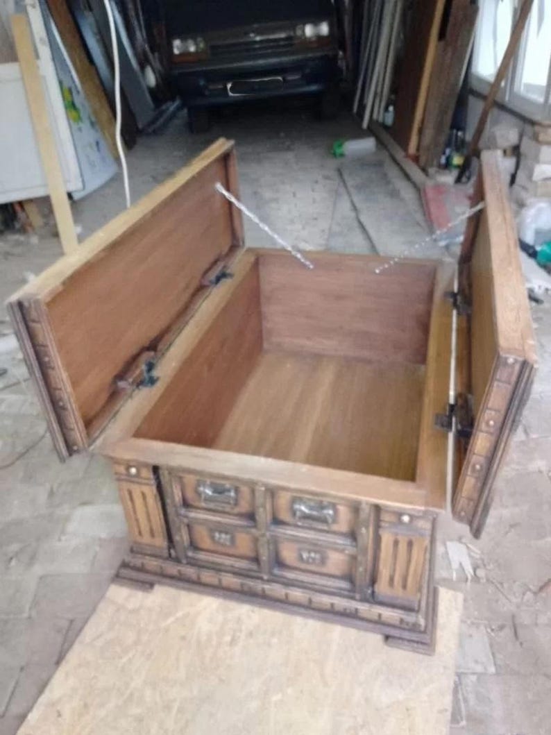 May include: A wooden chest with a hinged lid and three drawers. The chest is made of light brown wood with darker brown accents and metal hardware. The lid is open, revealing the interior of the chest.