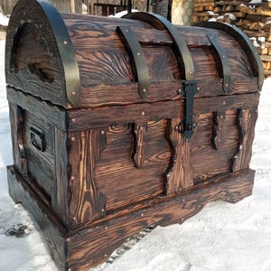 May include: A large, wooden chest with a dark brown finish. The chest features metal bands and a black latch. The wood grain is visible, and the chest has decorative carved details. The chest is a storage container.