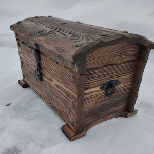 May include: A rustic wooden chest with a hinged lid, metal hardware, and a dark brown finish. The chest features decorative metal studs and a handle. The chest is elevated on small wooden feet.