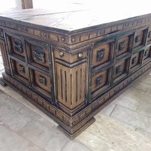 May include: A large, antique-style wooden chest with a dark brown finish. The chest features ornate carvings, metal accents, and decorative lion head handles. The design includes multiple square panels and a sturdy base, suggesting a substantial storage capacity.