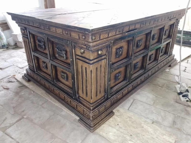 May include: A large, antique-style wooden chest with a dark brown finish. The chest features ornate carvings, metal accents, and decorative lion head handles. The design includes multiple square panels and a sturdy base, suggesting a substantial storage capacity.