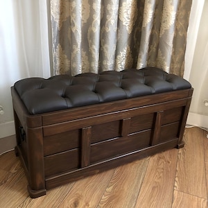 Pode incluir: Um baú de armazenamento de madeira castanha escura com uma almofada de couro castanho escuro almofadada. O baú tem uma forma retangular com painéis decorativos na frente e nas laterais. Está sobre um chão de madeira.