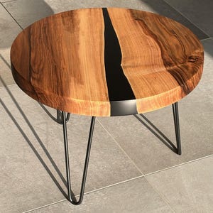 Op de afbeelding: Ronde houten salontafel met een donkere epoxyharsinleg en zwarte haarspeldpoten. Het tafelblad is gemaakt van natuurlijk hout met zichtbare nerf, en de epoxyhars zorgt voor een opvallend contrast. De tafel heeft een diameter van ongeveer 61 cm.