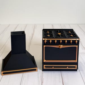 Pode incluir: Um conjunto de fogão e exaustor em miniatura pretos com acabamentos dourados. O fogão tem seis queimadores e uma gaveta. O exaustor tem formato retangular. O conjunto é para uma casa de bonecas ou cozinha em miniatura.