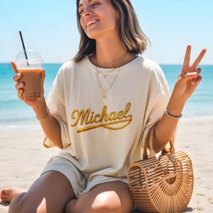 May include: Cream-colored t-shirt with the name "Michael" in gold script. The person is holding a clear plastic cup with a straw and a woven bamboo handbag. The background is a beach with blue water and sky.