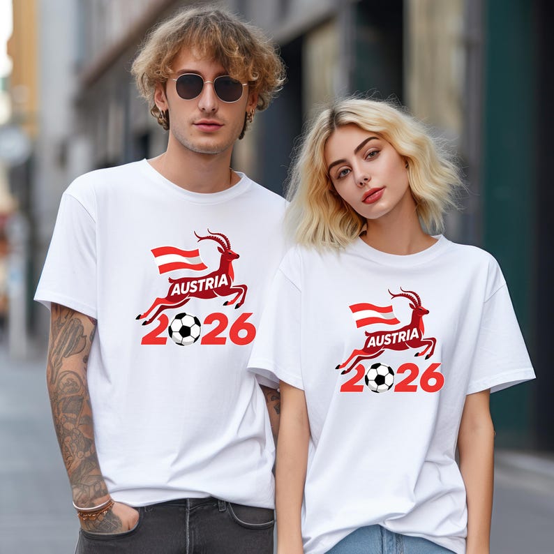 May include: Two people wearing white t-shirts with a red and black graphic. The graphic includes the word "AUSTRIA", a stylized deer, a soccer ball, and the number "2026". The Austrian flag is also featured in the design.