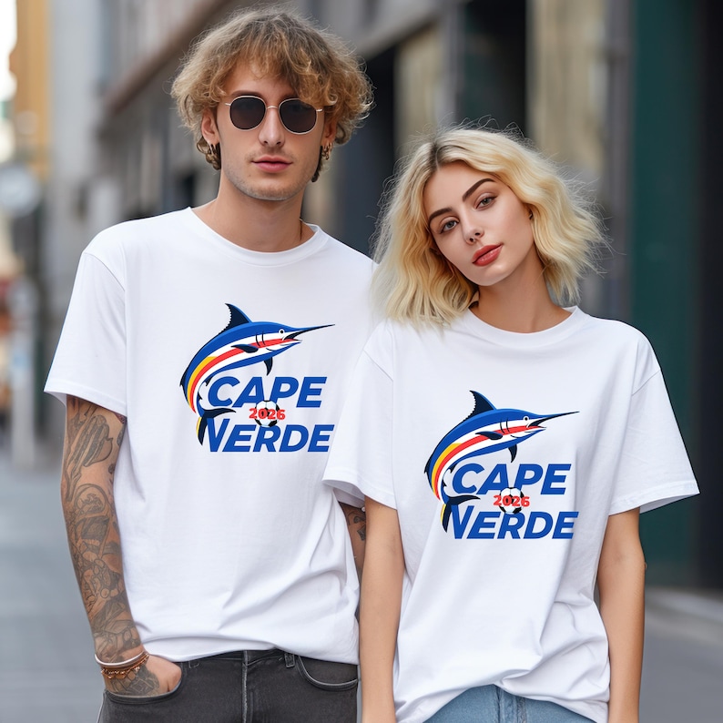 May include: Two people wearing white t-shirts with a blue marlin graphic and the words "CAPE VERDE 2026" in blue. The marlin has blue, red, and yellow accents. The background is a blurred city street.