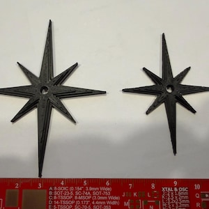 May include: Two black, eight-pointed star-shaped ornaments with a central circular hole. The larger star measures approximately 4.5 inches in height, and the smaller star measures approximately 3.5 inches in height, as shown by the red ruler.
