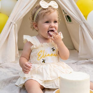 May include: A cream-colored ruffled romper featuring an embroidered bee design and the name Elianah in yellow script, paired with a matching cream bow headband. The garment includes a white number one graphic on the bodice.