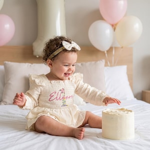 May include: A cream-colored long-sleeve romper with a smocked bodice, featuring the name Ellanor embroidered in pink script above a floral number one. The garment includes ruffled shoulder details and is paired with a matching cream-colored bow headband.