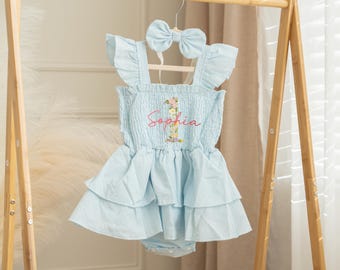 May include: A light blue baby dress with a matching bow headband, hanging on a wooden rack. The dress features a ruffled skirt and the name "Sophia" embroidered in pink with floral accents. The outfit is designed for a baby.