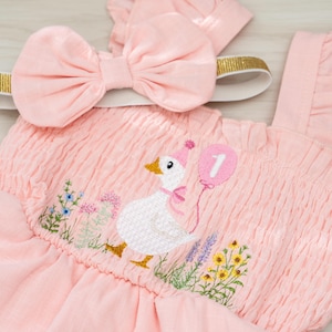 Op de afbeelding: Roze baby-outfit met bijpassende strik-hoofdband. De outfit heeft een geborduurde gans met een ballon met het nummer 1, omringd door bloemenborduurwerk. De hoofdband heeft een gouden glitterband.