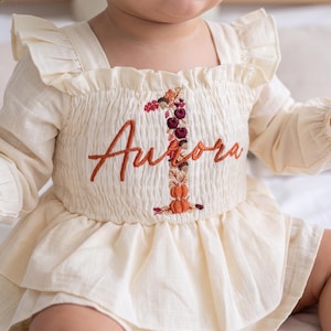 May include: A cream-colored long-sleeve dress with ruffled shoulders and a smocked bodice. The front features embroidered text reading Aurora in orange, overlaid on a large number one decorated with autumn-themed motifs like pumpkins and leaves.