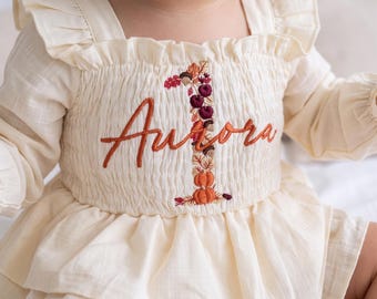 Könnte beinhalten: Ein cremefarbenes Langarmkleid mit gerüschten Schultern und gesmoktem Oberteil. Auf der Vorderseite befindet sich der in Orange gestickte Schriftzug Aurora, der über einer großen Eins liegt, die mit herbstlichen Motiven wie Kürbissen verziert ist.