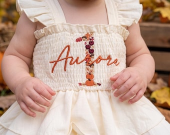 May include: A cream-colored sleeveless dress with ruffled shoulder straps and a smocked bodice. The front features embroidered text reading Aurora in rust-colored thread, positioned next to a vertical number one decorated with pumpkins and acorns.