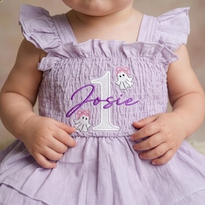 May include: A light purple smocked dress with ruffled shoulder straps. The front features an embroidered white number one, the name Josie in purple script, and two small white ghost figures with pink bows positioned above and below the name.