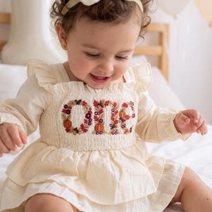 May include: A cream-colored long-sleeve dress with a smocked bodice featuring embroidered autumn-themed letters spelling out the word ONE, decorated with small pumpkins, acorns, and fall leaves in shades of orange, burgundy, and brown.