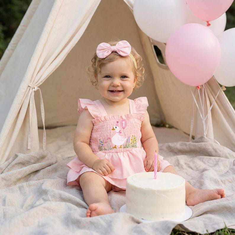 Op de afbeelding: Een lachend kind draagt een roze jurk met een eendenontwerp en een bijpassende haarband met strik. Voor hen staat een kleine taart met een brandende kaars, omringd door roze en witte ballonnen. Op de achtergrond staat een kleine tent.