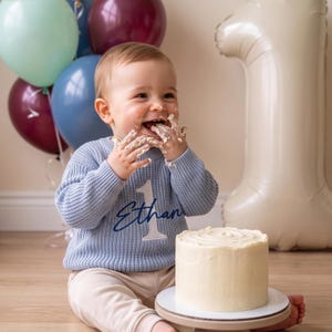 May include: A light blue long-sleeve knit sweater featuring a white embroidered number one and the name Ethan in dark blue script. The sweater is worn with beige pants, and the garment is clean with a textured waffle-knit pattern.