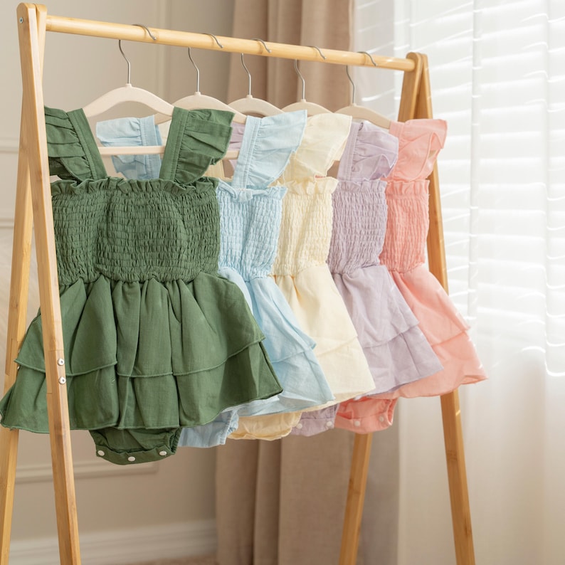 Op de afbeelding: Een rij babyjurkjes in verschillende pastelkleuren, waaronder groen, blauw, geel, paars en roze, hangend aan een houten rek. Elke jurk heeft een gerimpeld lijfje en een gelaagde rok, perfect voor de babygarderobe.