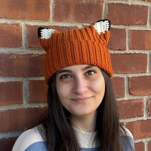May include: An orange knitted beanie hat with fox ears. The hat has a ribbed design and is made of a warm, textured material. The fox ears are white with black tips. A playful accessory.