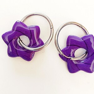 Puede incluir: Un par de pendientes morados en forma de estrella con aros plateados. Los pendientes tienen un acabado brillante y un patrón de remolino morado oscuro. Sobre un fondo blanco.