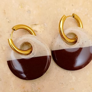 Peut inclure: Une paire de boucles d'oreilles créoles composées d'anneaux en métal doré et de pendentifs circulaires. Chaque pendentif est divisé en deux parties, avec une section supérieure crème mouchetée et une section inférieure marron foncé brillant.